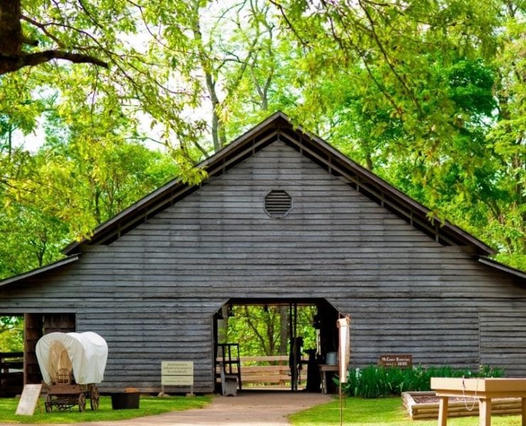 A model covered wagon is parked outside a replica 1800s barn for Burritt's Spring Farm Days event.