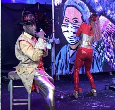 a man sitting on a stage at the ArtRageous show painting a portrait of a nurse wearing a face mask