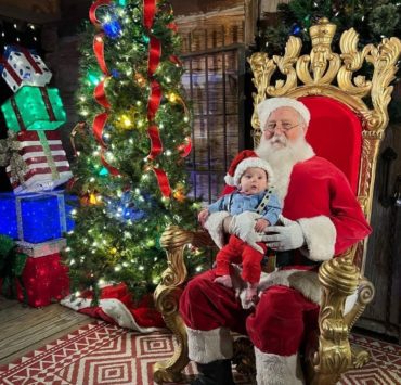 A baby in a blue button down shirt with Christmas tree suspenders and a knit Santa hat sits in Santa's lap at Christmas at the Falls.