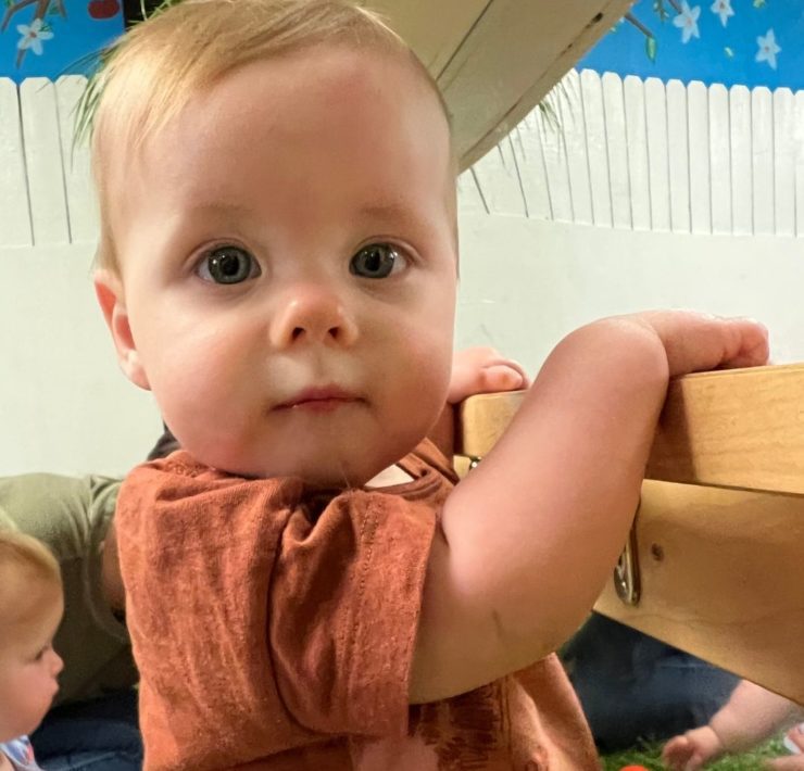 A baby boy stands with the assistance of a pull up bar that's placed in front of a mirror.
