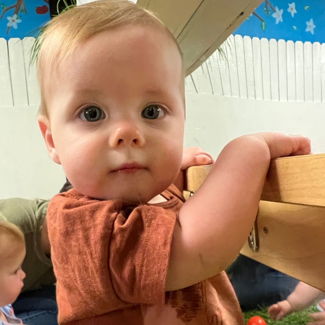 A baby boy stands with the assistance of a pull up bar that's placed in front of a mirror.