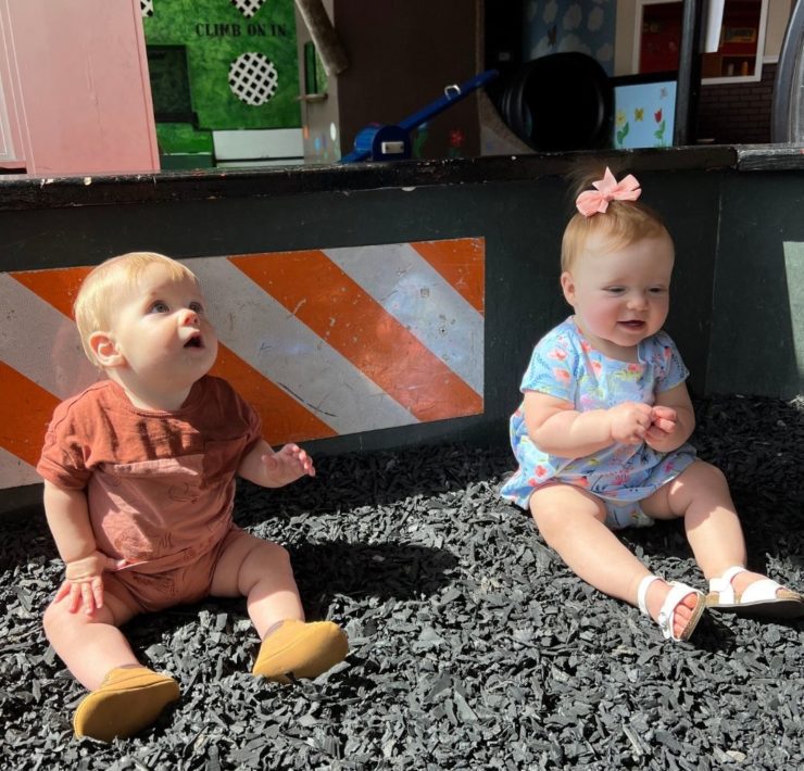Two babies sit in black rubber mulch in the construction zone at the Imagination Place.