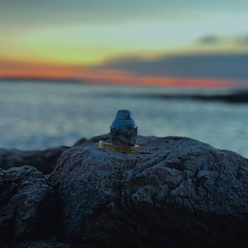 A small urn rests on a rocky cliffside overlooking the ocean as the sun sets in the background. 