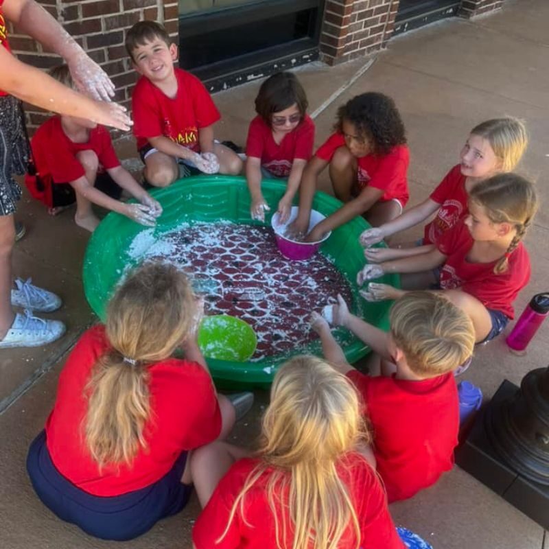 A group of campers wearing red Kidcam Camps shirts sit in a circle around a green kiddie pool during a craft.