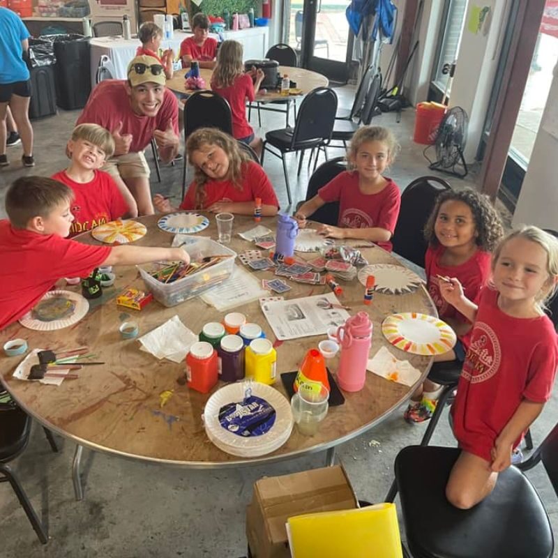 A group of campers in red shirts sit around a wooden table with painting supplies sprawled across the face of it.