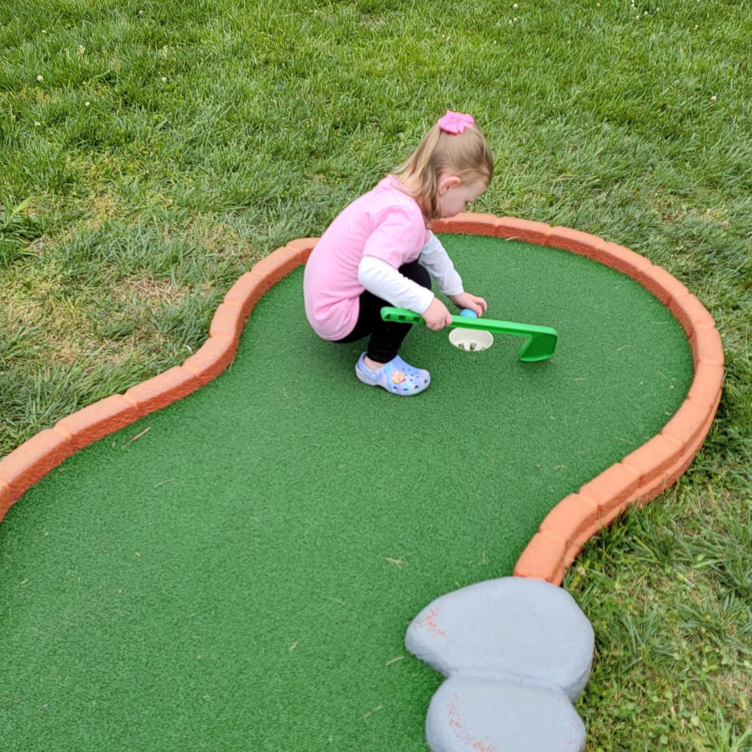A toddler girl in pink crouches on a mini golf course hole with a plastic green club in hand.