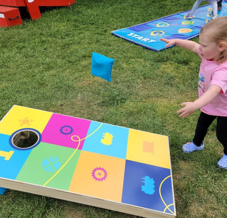 A young girl tosses a blue bean bag towards a colorful cornhole board.