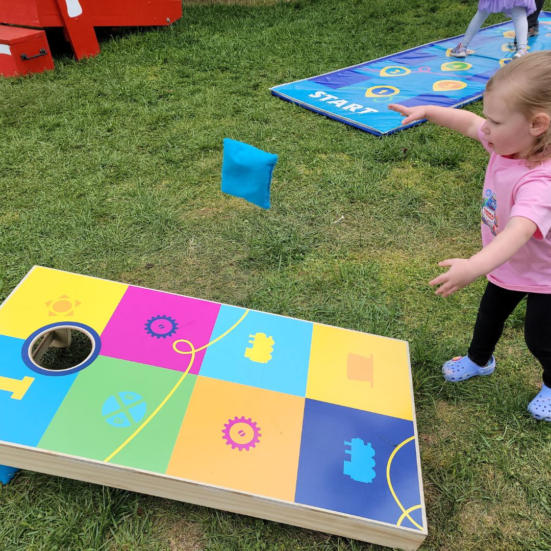A young girl tosses a blue bean bag towards a colorful cornhole board.