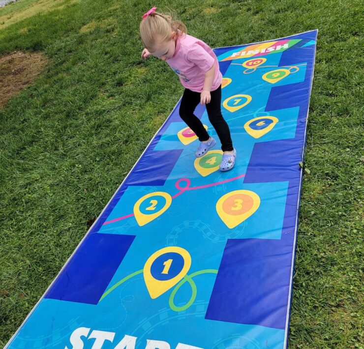 A toddler girl in pink hops her way down a blue checked hopscotch mat.