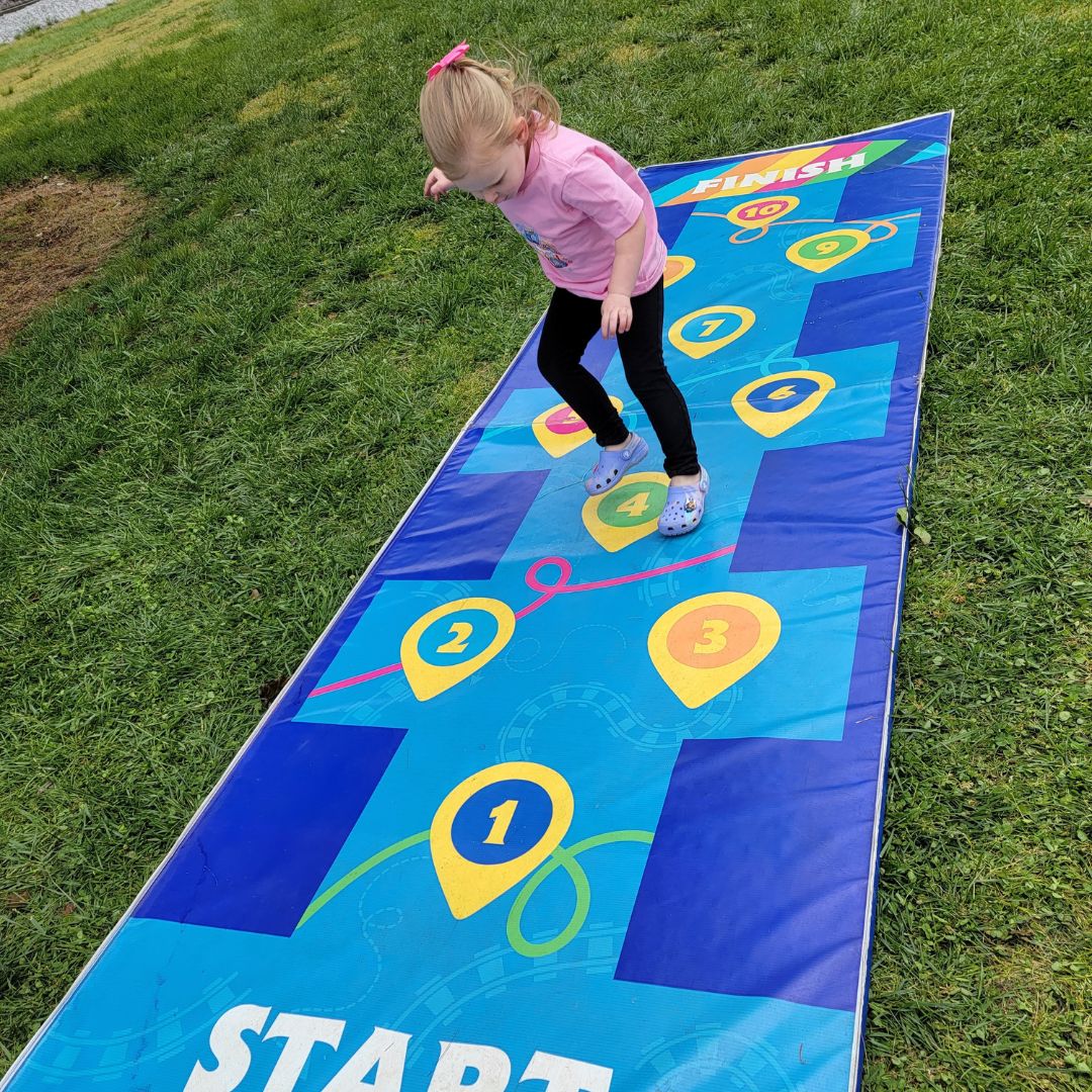 A toddler girl in pink hops her way down a blue checked hopscotch mat.