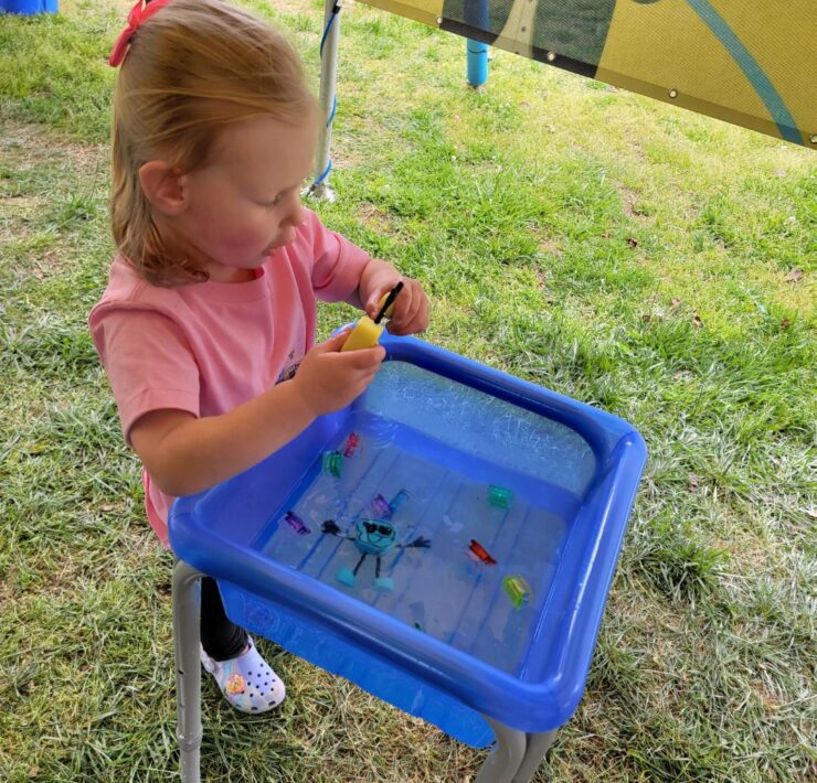A toddler girl in pink plays at a small blue water table with small character toys inside it.