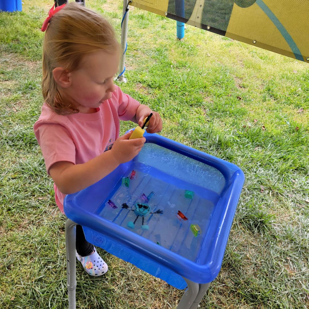 A toddler girl in pink plays at a small blue water table with small character toys inside it.