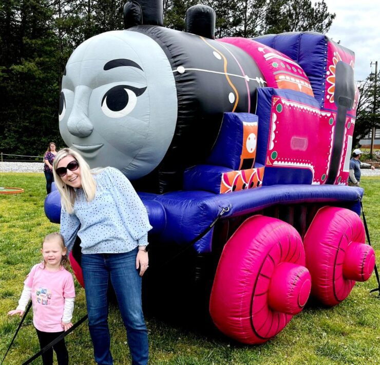 A toddler girl and pink and her mother stand in front of a pink Thomas the Train character.