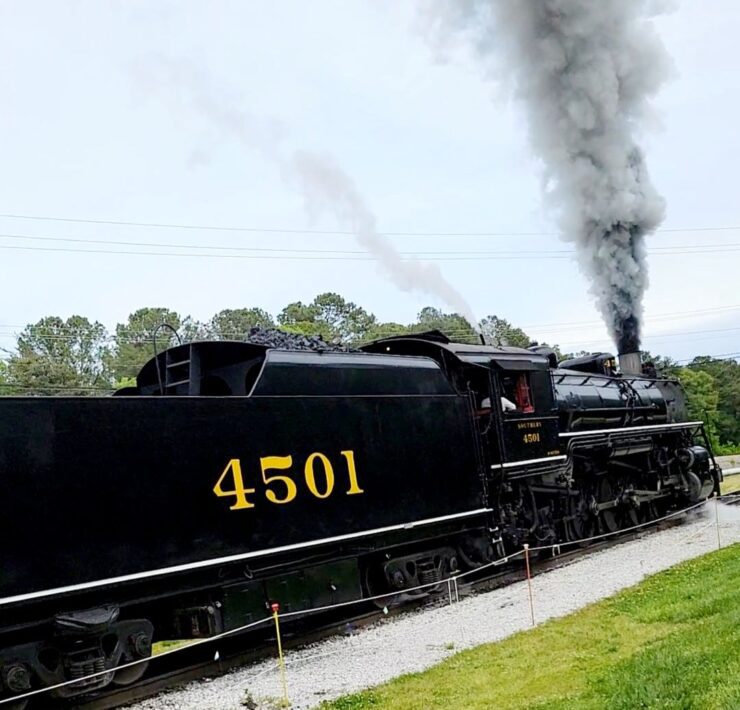 A black train engine with smoke billowing from it makes its way down the railroad tracks at the Tennessee Valley Railroad Museum.
