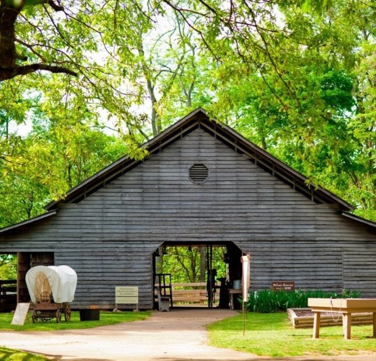 A model covered wagon is parked outside a replica 1800s barn for Burritt's Spring Farm Days event.
