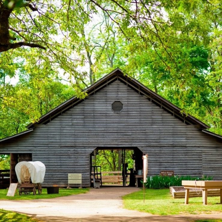 A model covered wagon is parked outside a replica 1800s barn for Burritt's Spring Farm Days event.