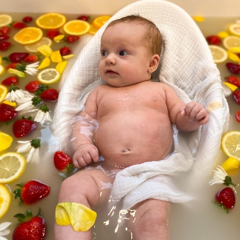 A baby lies in a white swaddle draped bath seat surrounded by a strawberry and lemon milk bath for this DIY photoshoot.