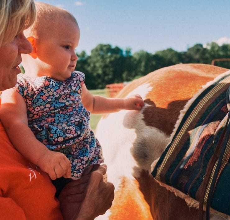 A toddler wearing a floral dress pets a brown and white horse with the help of LeAnn Newsom at Haven Farms.