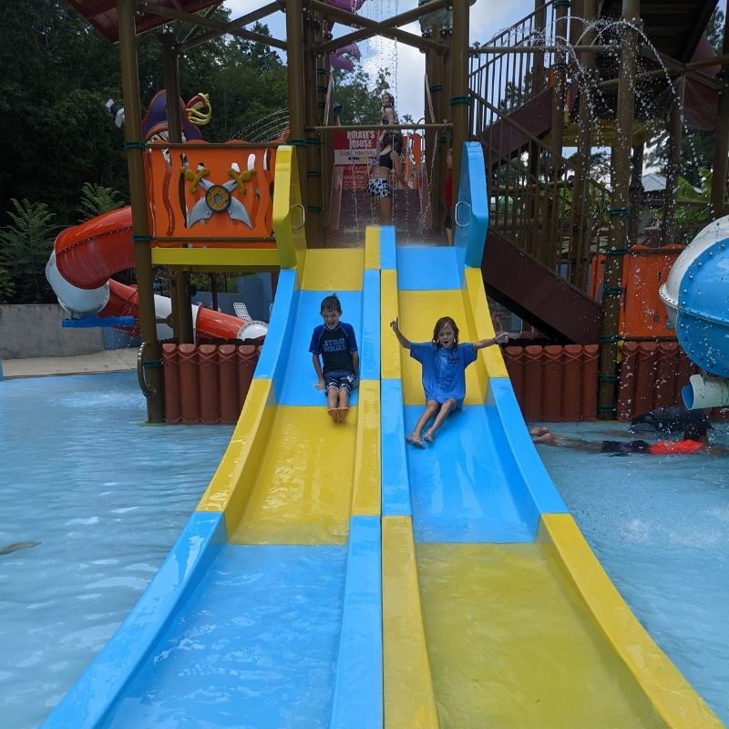 Two kids slide down twin blue and green slides from a pirate ship shaped play area.