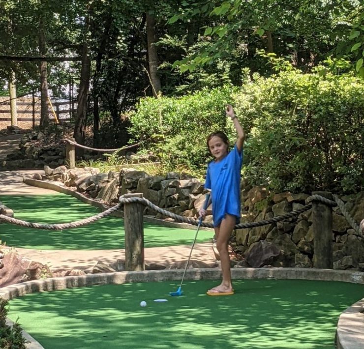 A young girl waves to the camera as she holds a putter for mini golf.