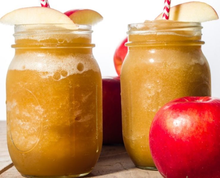 Two apple slushies sit on a wooden background with apple slice garnishes and a red and white striped straw.