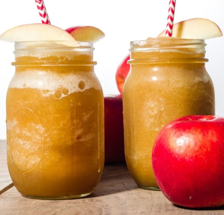 Two apple slushies sit on a wooden background with apple slice garnishes and a red and white striped straw.