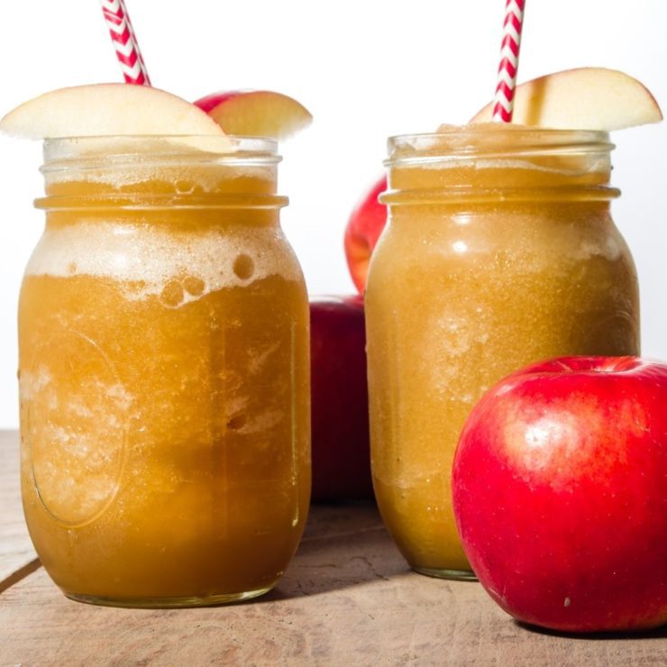 Two apple slushies sit on a wooden background with apple slice garnishes and a red and white striped straw.