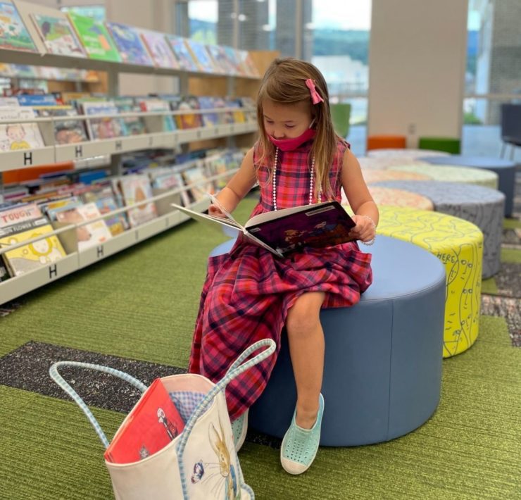 A young girl in a pink plaid dress reads as she sits on a pale blue stool in the children's area of the South Huntsville Library.