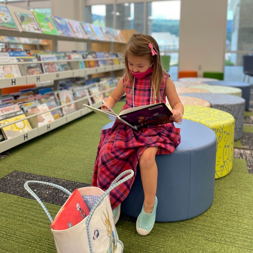 A young girl in a pink plaid dress reads as she sits on a pale blue stool in the children's area of the South Huntsville Library.