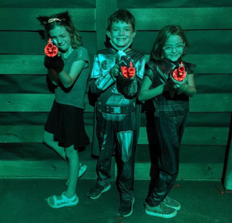 Three kids hold toy dart guns towards the camera in a green lit room.