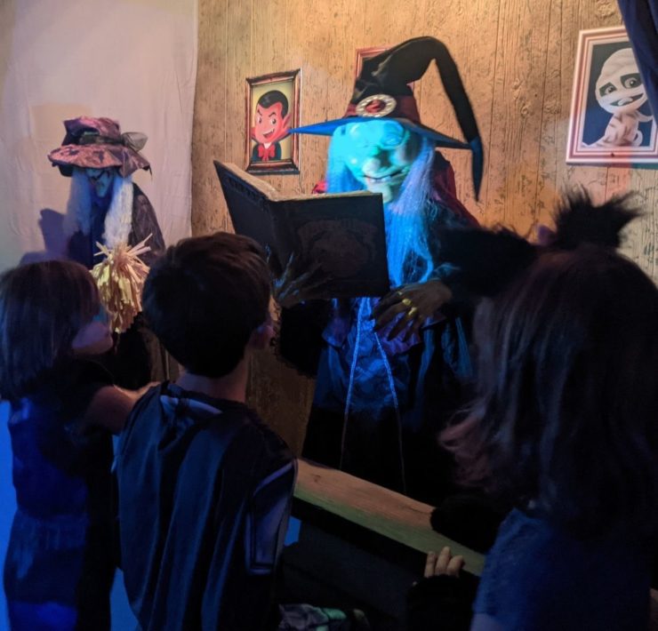 Three children peer over a ledge at an illuminated witch holding a book.