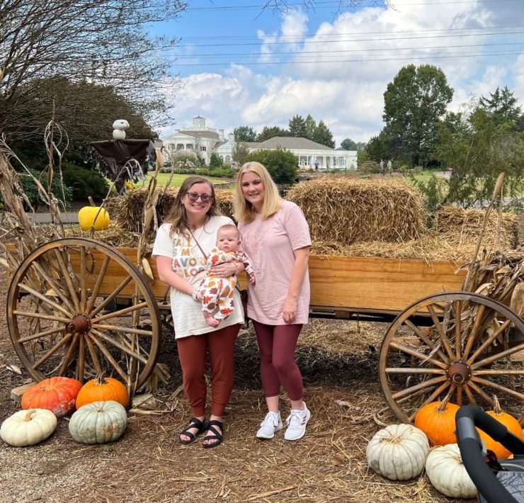 A mom and grandmother hold a baby in front of a trailer full of hay and pumpkins during their Scarecrow Day at the Huntsville Botanical Garden.