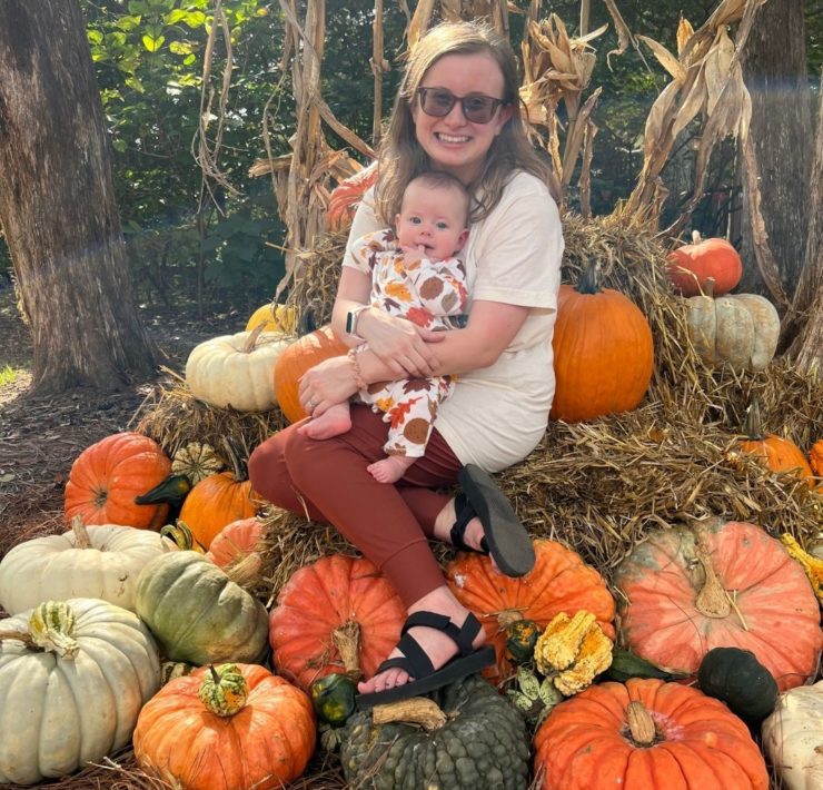 A mom holds her baby while sitting on a a hay bale surrounded by pumpkins during their Scarecrow Day at the Huntsville Botanical Garden.