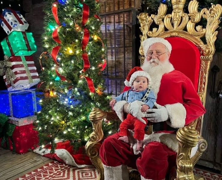 A baby in a blue button down shirt with Christmas tree suspenders and a knit Santa hat sits in Santa's lap at Christmas at the Falls.