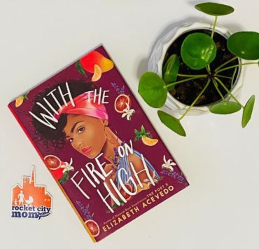 The book "With the Fire on High" sits on a white table next to a green plant and orange and purple Rocket City Mom sticker.