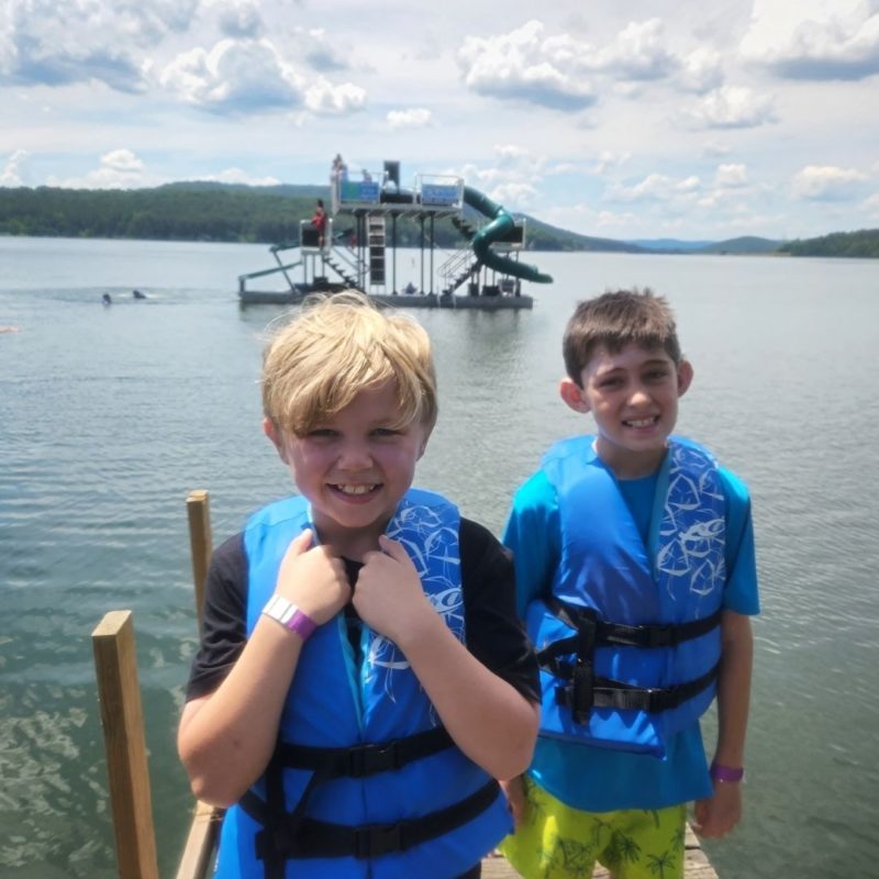 Two boys in blue life jackets stand on the dock in front of the floating adventure course, The Jungle Wave.