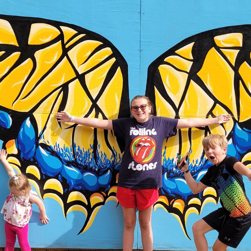 Three children of varying ages pose in front of a yellow and blue butterfly mural at Callaway Gardens.