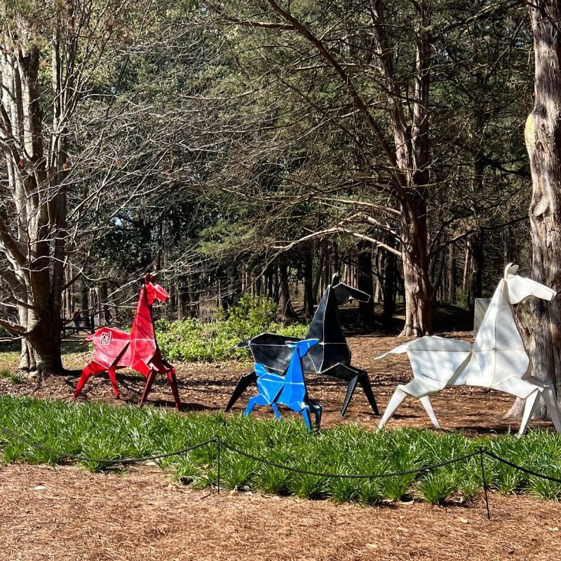 A red, blue, black, and white origami horse "gallop" through the trees at the Huntsville Botanical Garden.