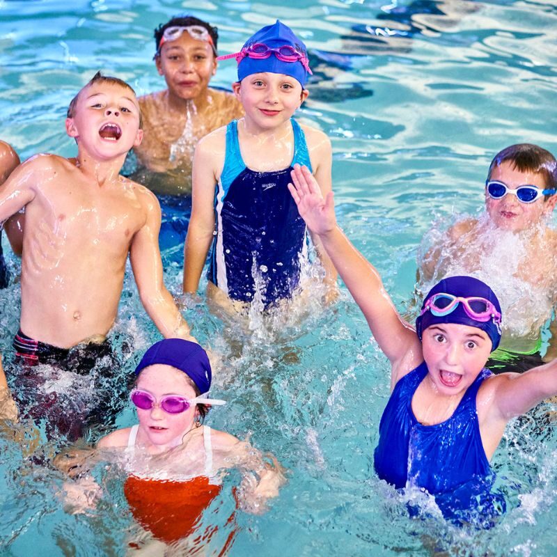 Children in bright bathing suits and goggles are splashing around during their swim lessons at SafeSplash Swim School.