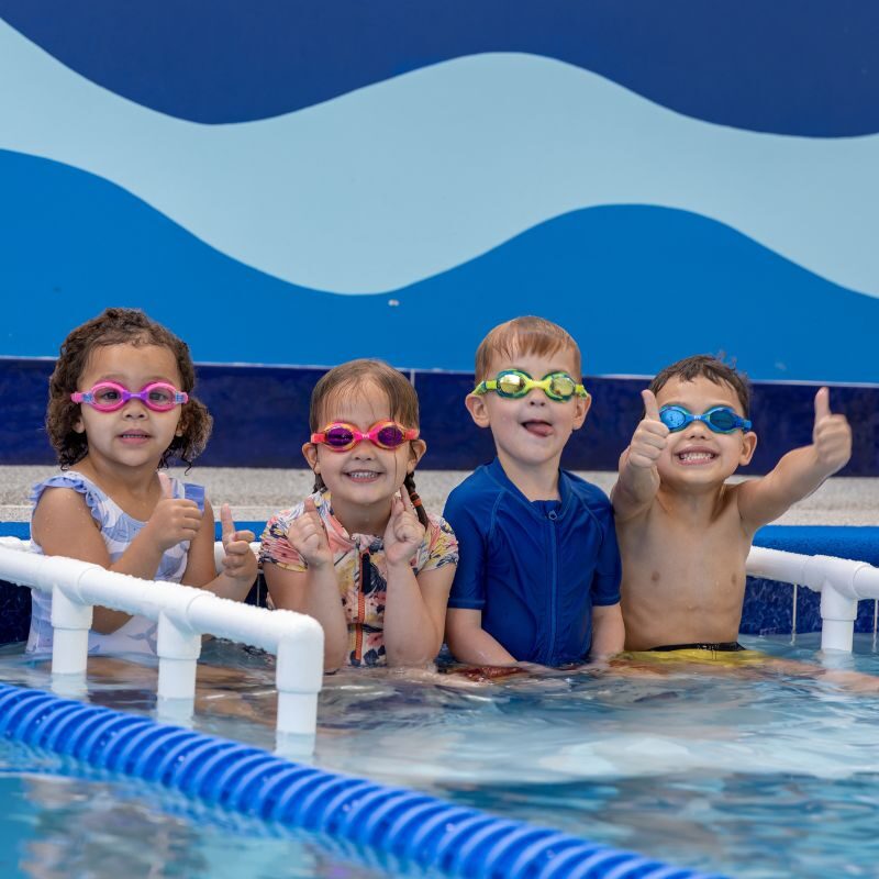 Four children in swimsuits and goggles sit on the edge of the pool before their swim lessons begin.