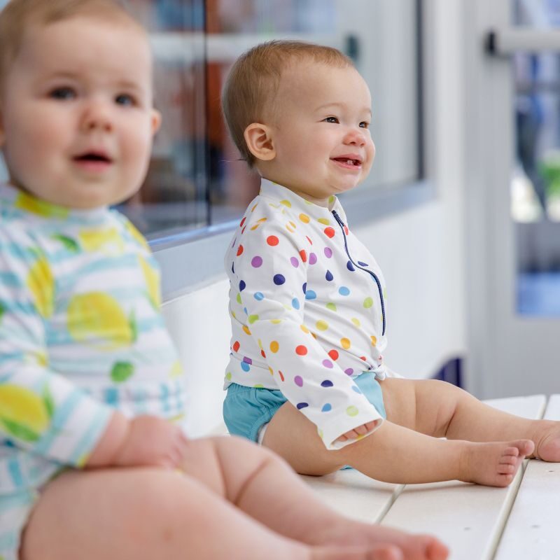 Two smiling babies sit on a white bench in their colorful swim suits waiting on their swim lessons to begin.