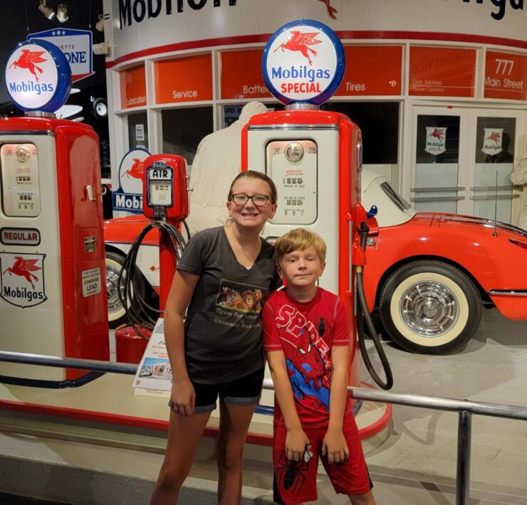 A young girl and her brother stand in front of vintage red and white gas pumps and a corvette at the National Corvette Museum.