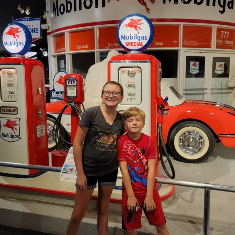 A young girl and her brother stand in front of vintage red and white gas pumps and a corvette at the National Corvette Museum. 