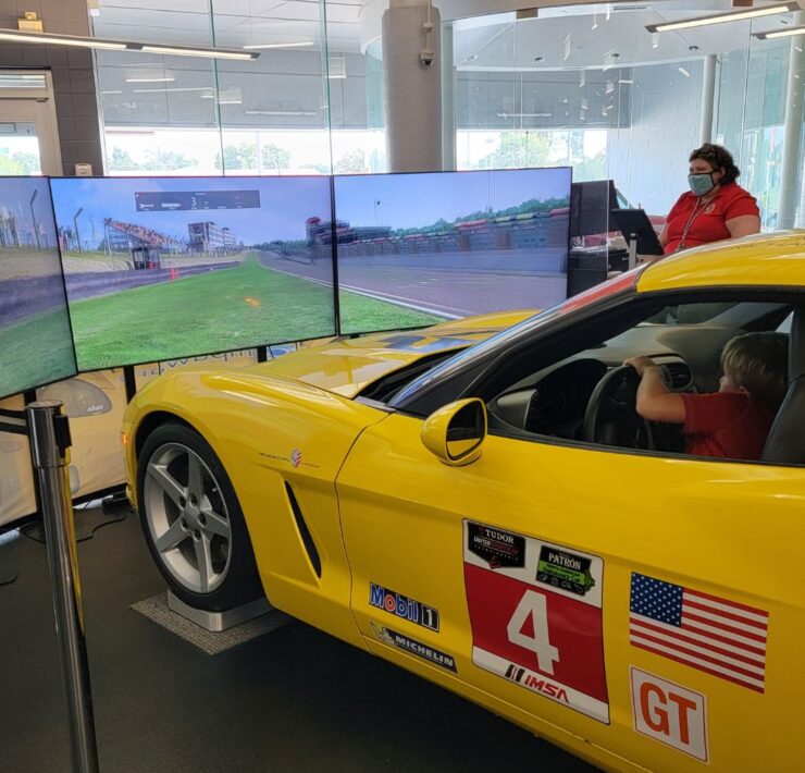 A bright yellow corvette sits behind three large plasma screen tvs as part of a corvette simulator at the National Corvette Museum.