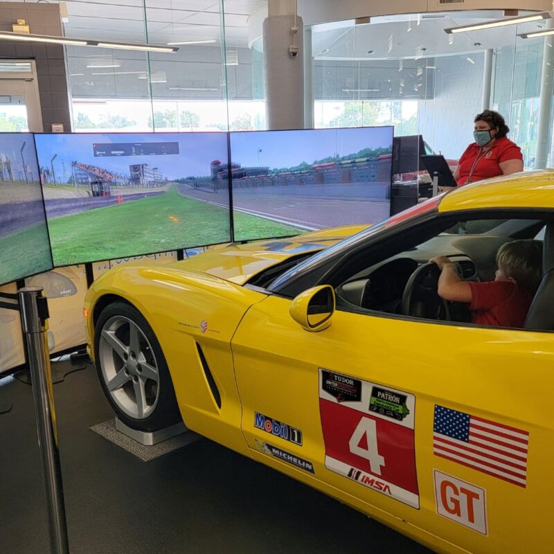 A bright yellow corvette sits behind three large plasma screen tvs as part of a corvette simulator at the National Corvette Museum.