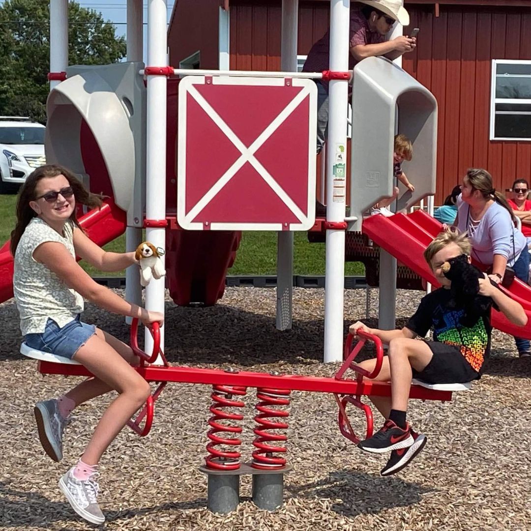 A young girl and boy play on a red seesaw with a barn themed playground behind them at Chaney's Dairy Barn in Bowling Green, KY.