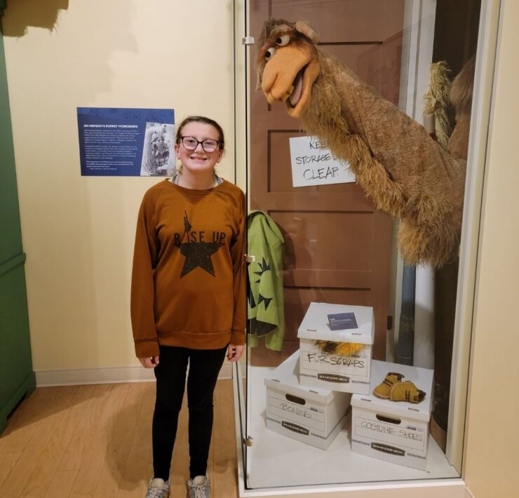 A teenage girl in a brown hoodie stands next to Sopwith the Camel from The Muppets.