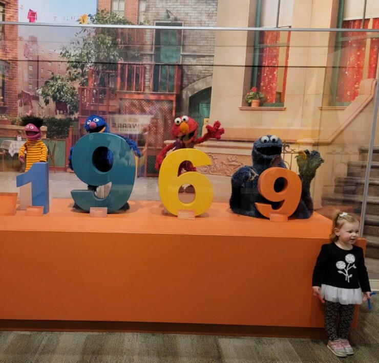 A young girl stands next to a display case with Sesame Street characters peeping around the numbers of 1969.