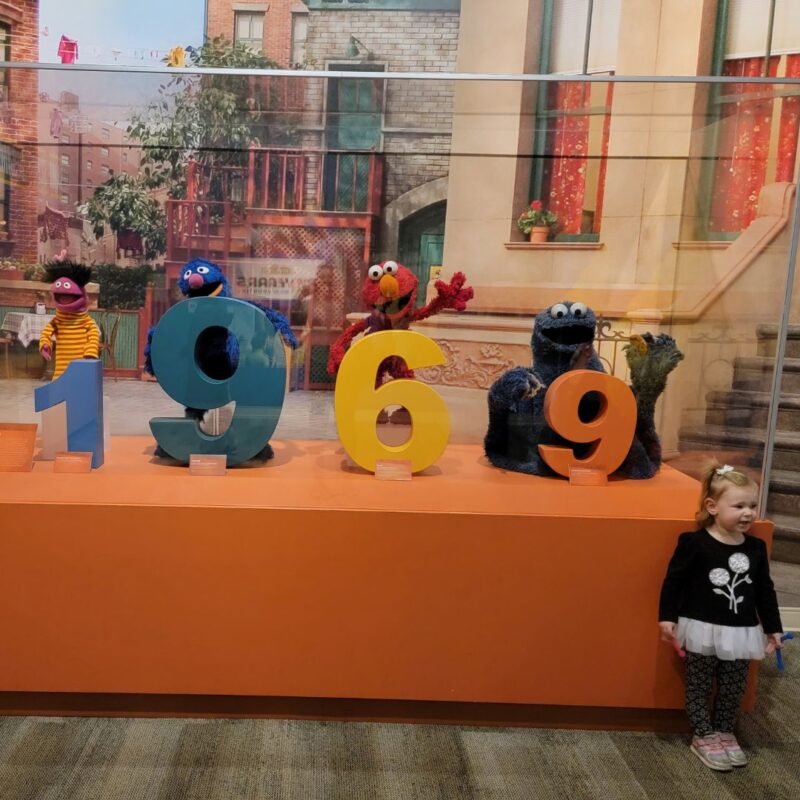 A young girl stands next to a display case with Sesame Street characters peeping around the numbers of 1969.