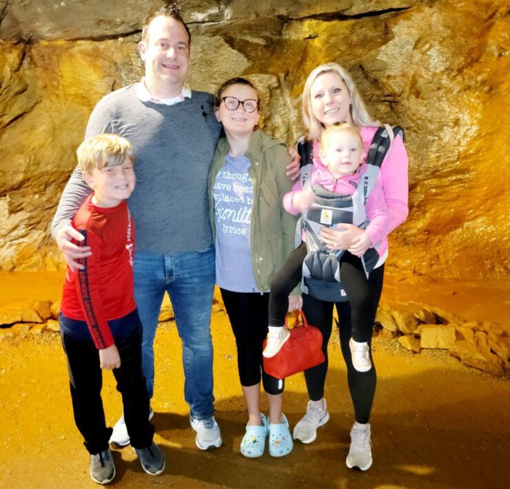 A family including a husband and wife, preteen girl, school-aged boy, and toddler girl in a baby carrier pose in front of rock walls in the Consolidated Gold Mine.